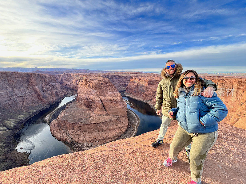 一对年轻的冒险旅行者拉丁异性恋夫妇在美国亚利桑那州格伦/大峡谷国家公园的马蹄形弯道顶部休息时欣赏风景，在冬季日落时分。